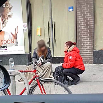 A woman in red jacket sitting on the sidewalk next to a bicycle.